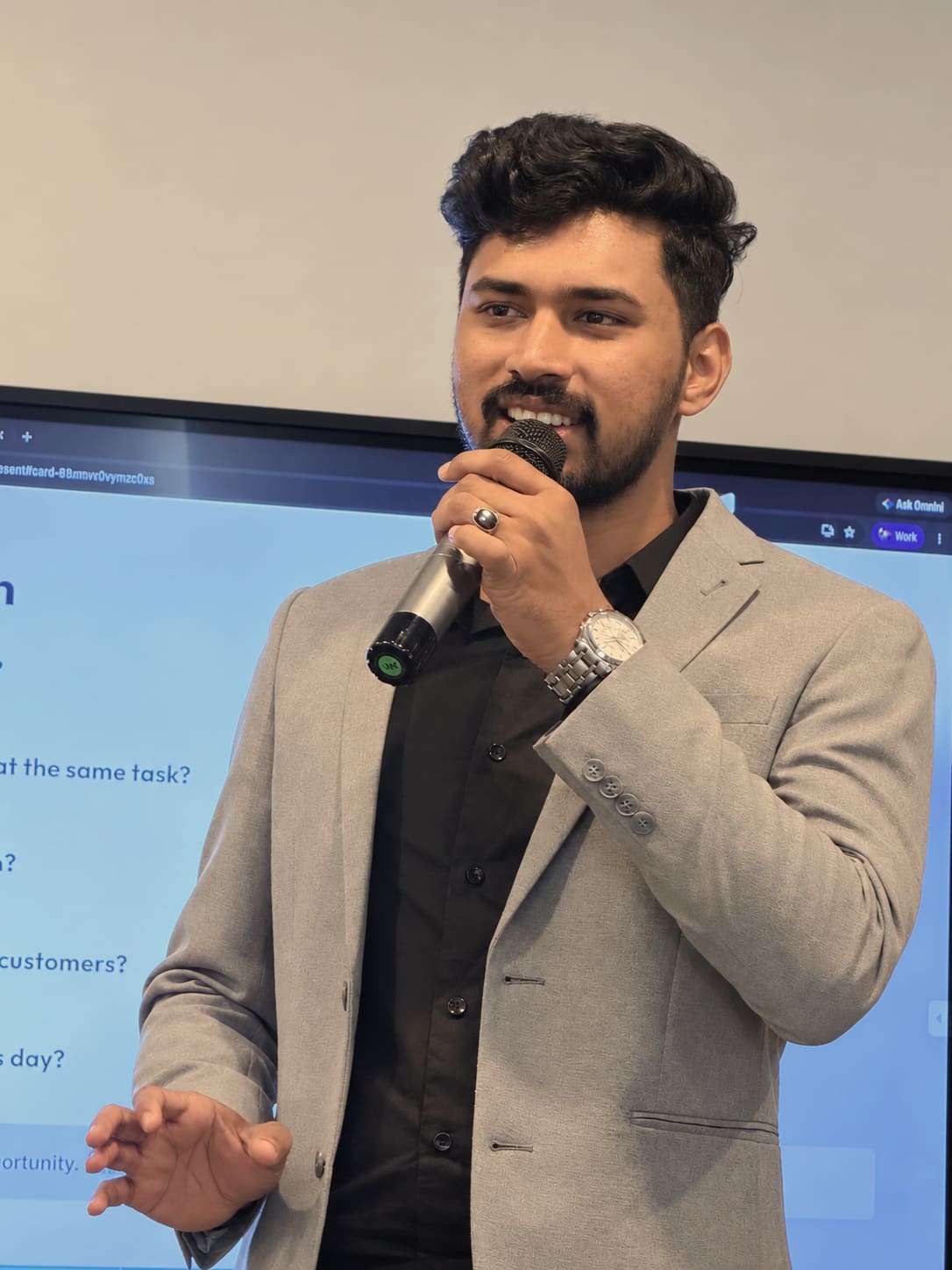 A smiling trainer in a grey blazer speaks into a microphone in front of a presentation screen